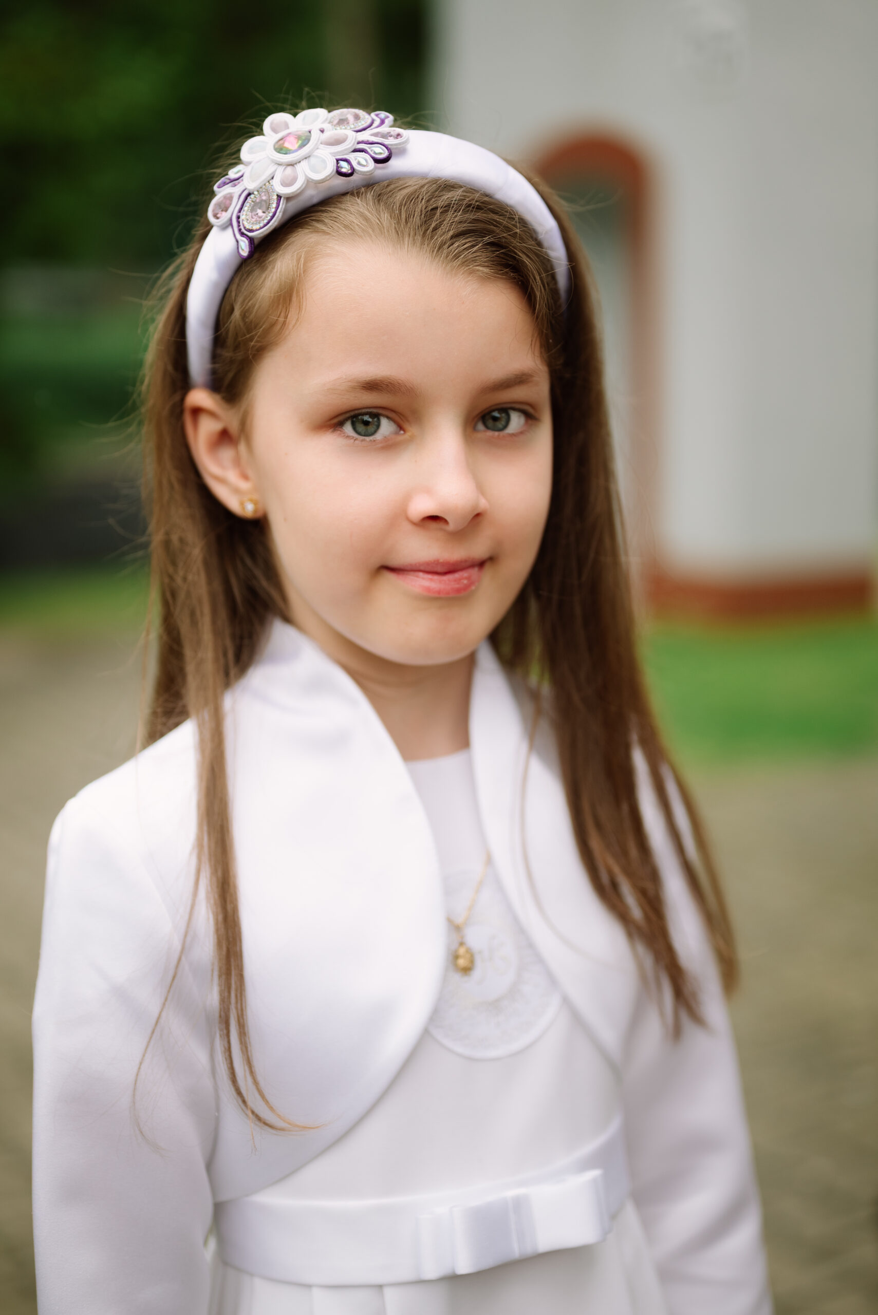 Portret dziewczynki w białym stroju komunijnym i eleganckiej, ręcznie robionej opasce sutasz z fioletowymi i różowymi kryształami. Ekskluzywna ozdoba komunijna od PHOENIX by MM.