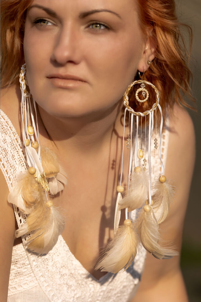 Ręcznie wykonane modularne kolczyki boho 2w1 z pozłacanej stali chirurgicznej PHOENIX by MM z odpinanymi piórami w kolorach bieli, kremu i beżu, zaprezentowane na modelce w stylizacji festiwalowej.